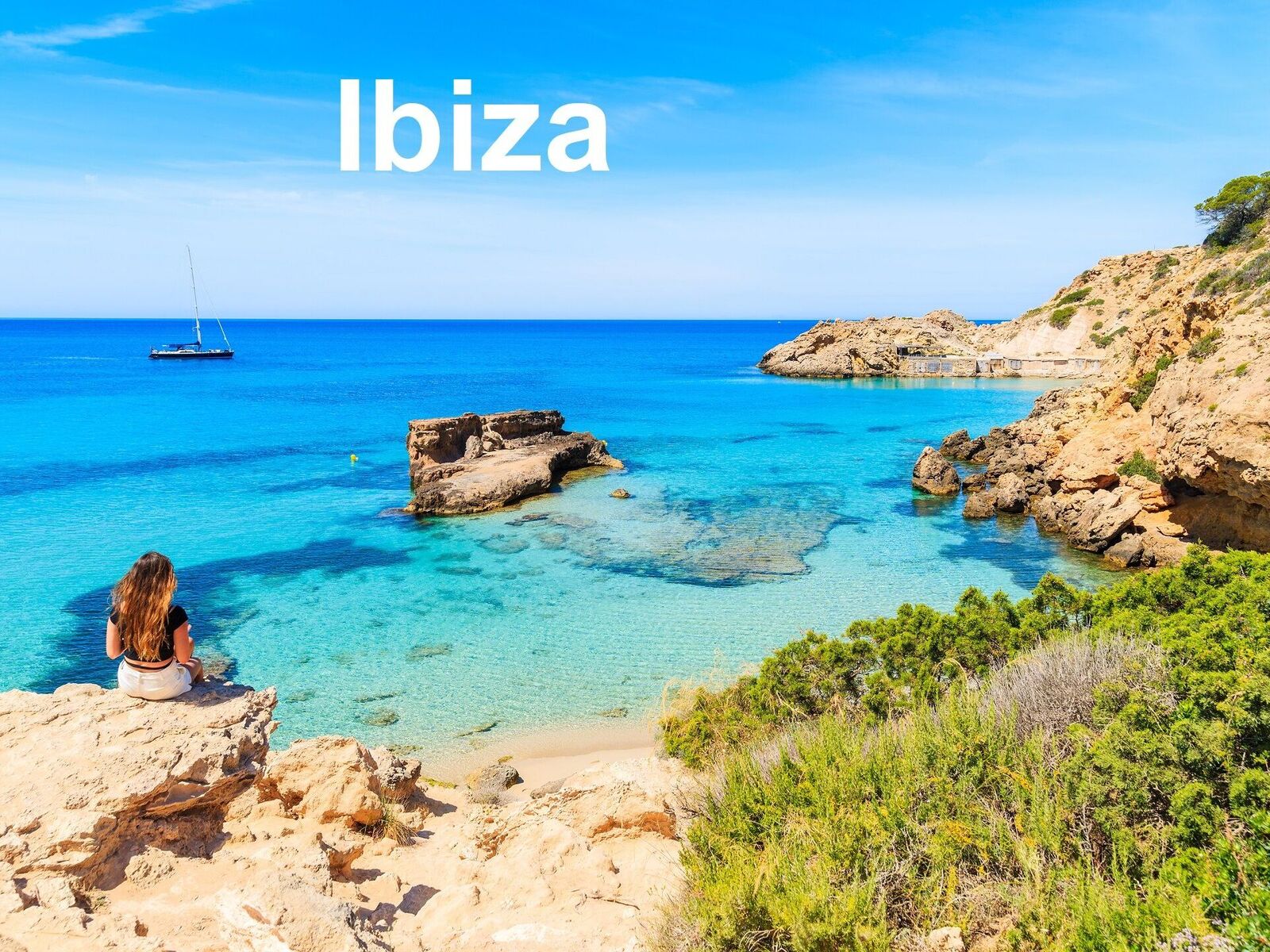 Unidentified young woman sitting on a rock and looking at beautiful Cala Tarida bay, Ibiza island, Spain